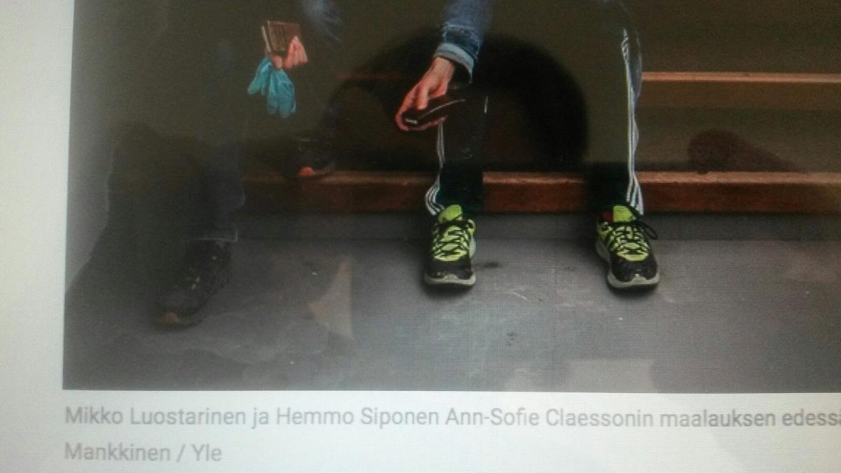 Kuva lehtileikkeleestä, jossa näkyy kuva ja kuvateksti. Kuva esittää kahden portailla istuvan ihmisen jalkoja, joissa on lenkkarit. Ihmisten käsissä on esineitä, mahdollisesti kumihanskat ja kännykkä. Kuvatekstinä lukee "Mikko Luostarinen ja Hemmo Siponen Ann-Sofie Claessonin maalauksen edessä. Mankkinen / Yle".