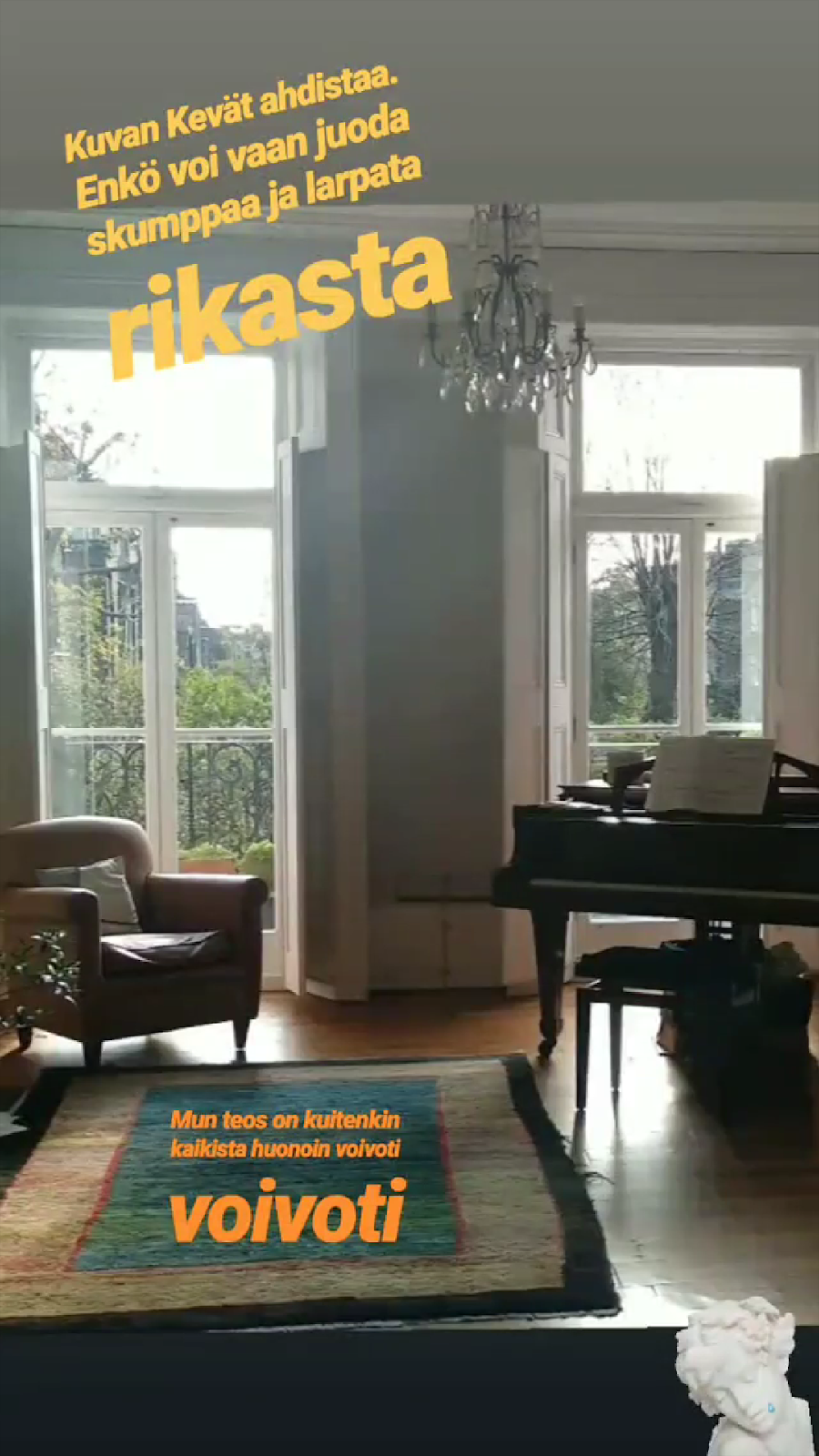 Olohuone, jossa kaksi suurta, korkeaa ikkunaa. Etualalla matto, katossa kristallikruunu ja ikkunoiden edessä vasemmalla ruskea nahkanojatuoli ja oikealla musta piano. Kuvaan lisätty oranssilla yläosaan teksti "Kuvan Kevät ahdistaa. Enkö voi vaan juoda skumppaa ja larpata rikasta" ja alaosaan teksti "Mun teos on kuitenkin kaikista huonoin voivoti voivoti"