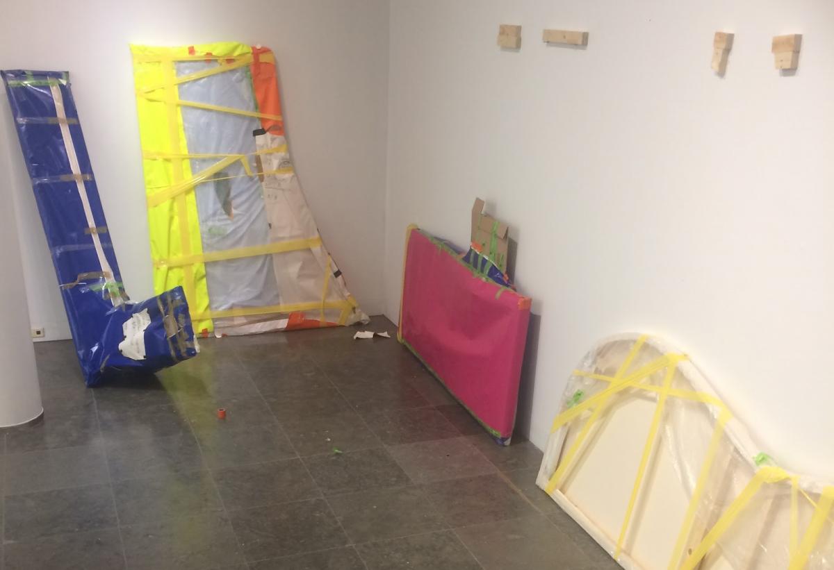 Valkoseinäoisen huoneen nurkka. Seiniä vasten nojaa paketteihin käärittyjä parin metrin kokoisia erimuotoisia levyjä. Levyt on kääritty esimerkiksi sinisiin, pinkkeihin, valkoisiin ja keltaisiin kääreisiin.