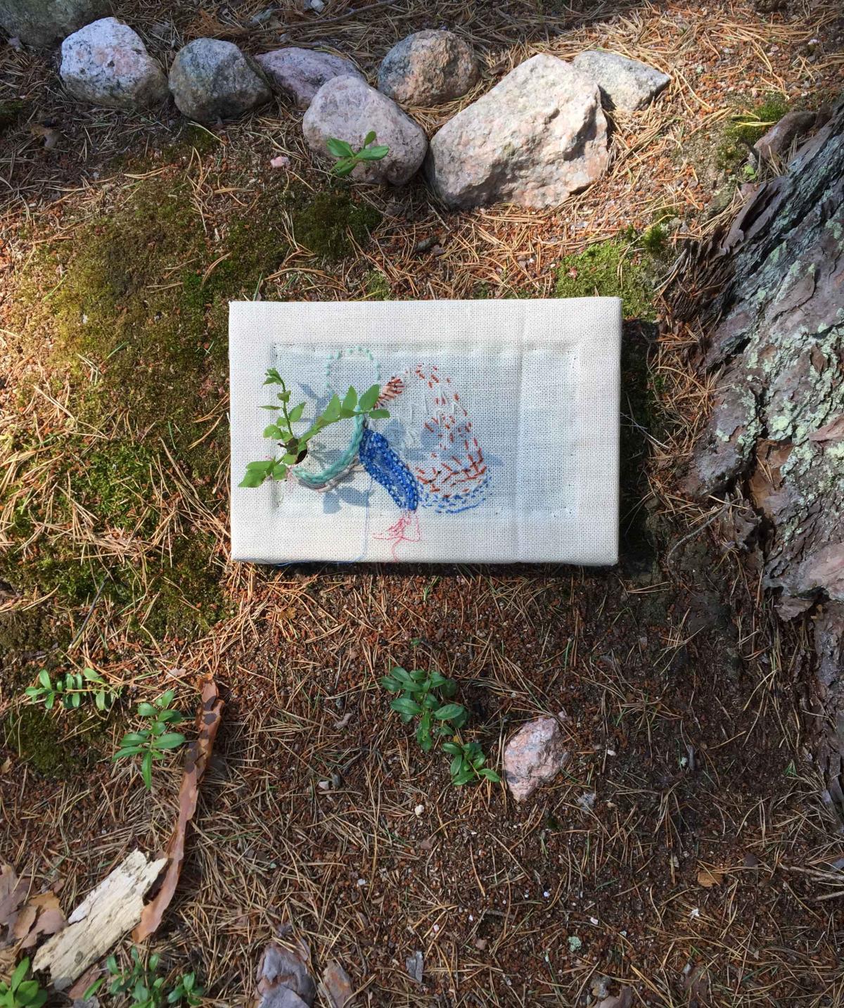 Kirjontateos maassa. Teoksen reiästä on pujotettu mustikan varpu.