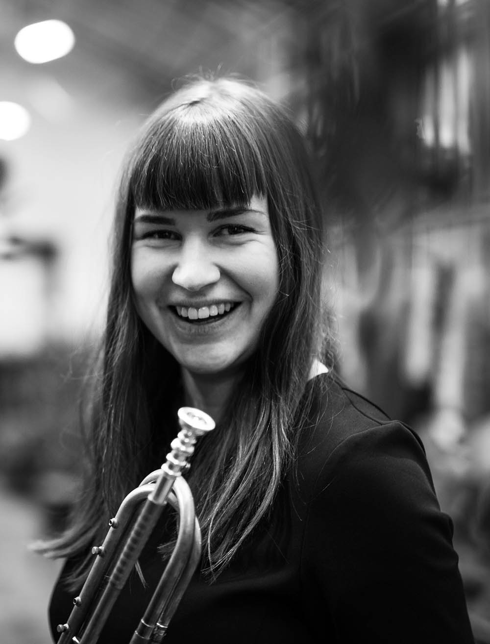 Inka Pärssinen hymyilee kuvassa kameralle. Kuva on mustavalkoinen ja hänellä on puhallinsoitin käsissään.