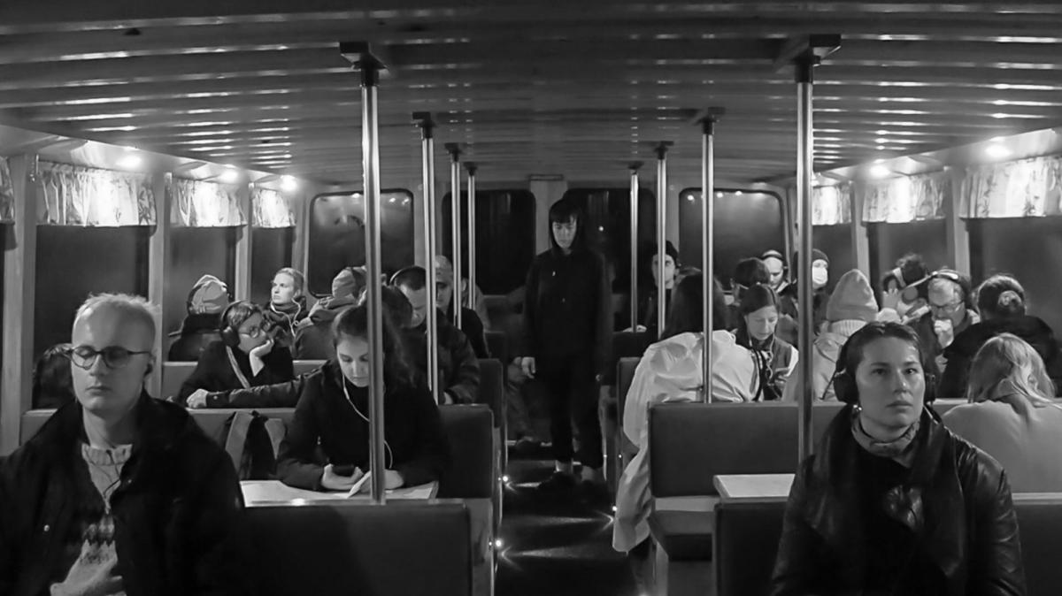In the photo, Minerva Juolahti walks on the aisle of the inside of a boat while performing the piece remembering by translating. Audience is sitting around tables and listening to the sound piece that is part of the performance. 