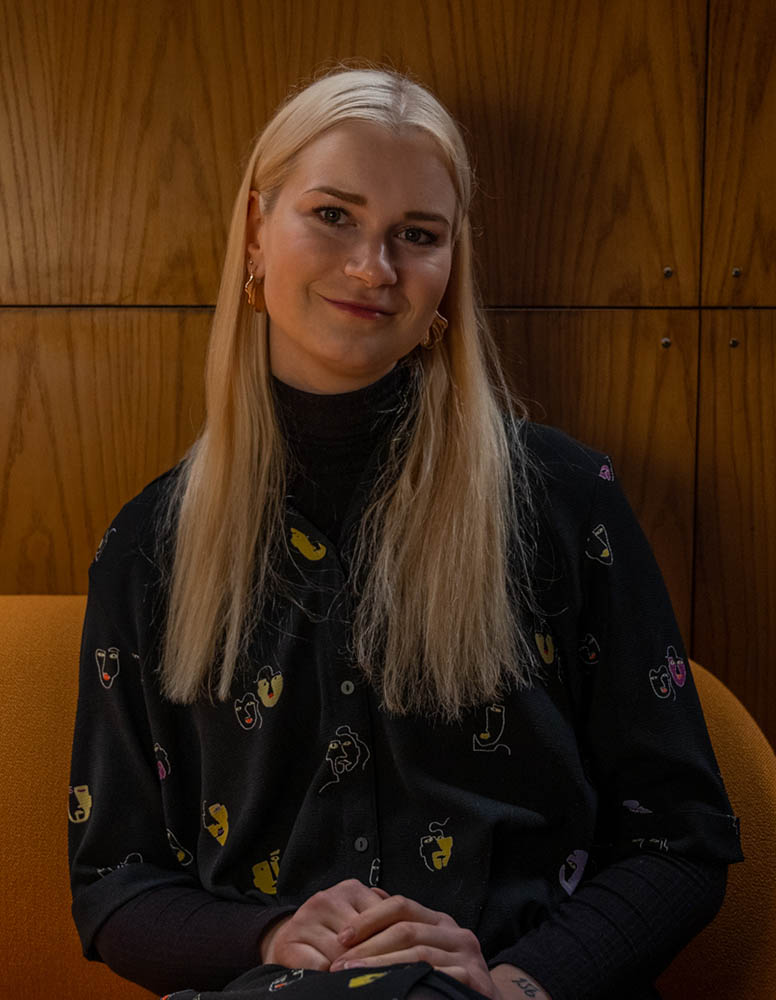 Meri Jaakola istuu puisen seinän edessä ja hymyilee kameralle.