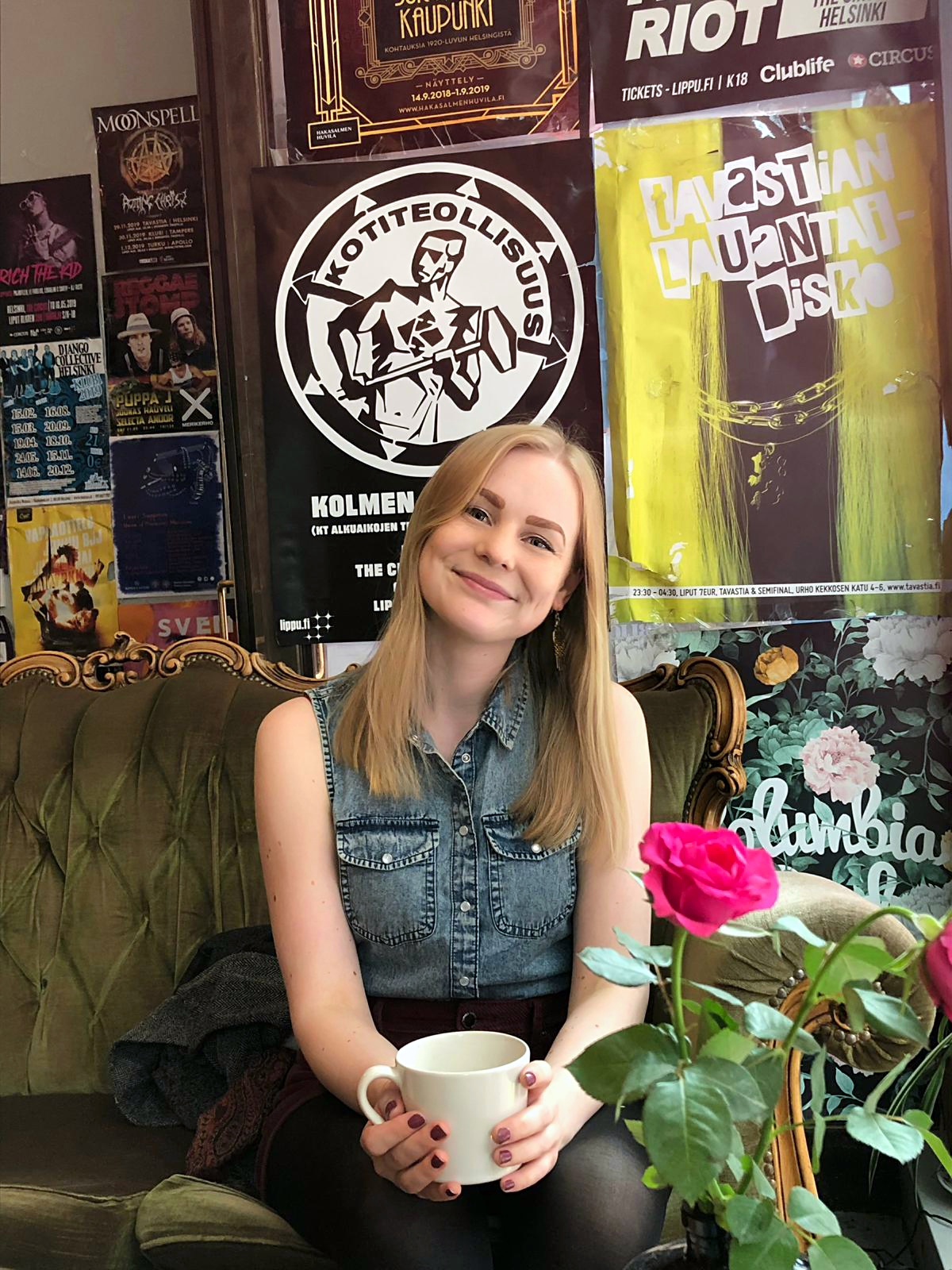 Linda Kanninen istuu kahvilassa kahvikuppi käsissään ja hymyilee kameralle. Taustalla on paljon julisteita ja sohva, jolla Linda istuu, on pramea ja viktoriaaninen.