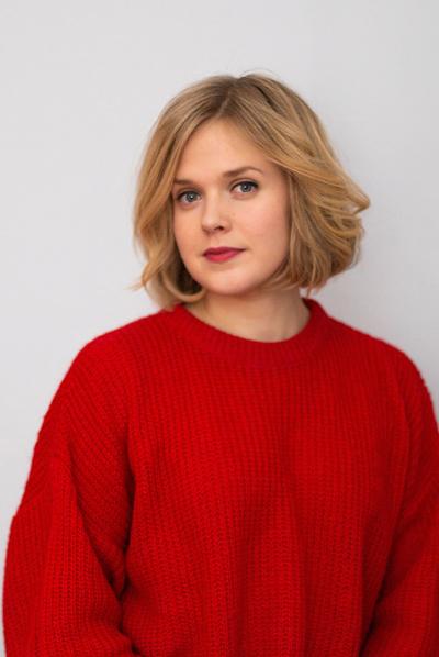 Suvi Ahoniemi on valkoisen seinän edessä. Päällä hänellä on punainen neule. Hän katsoo kameraan neutraali ilme kasvoillaan.